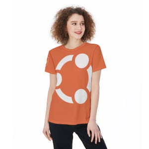 Peut inclure: Un t-shirt orange avec un grand motif graphique blanc. Le t-shirt a des manches courtes et un col rond. Le motif est un logo circulaire stylisé. La personne porte un pantalon noir.