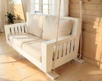 May include: White wooden swing with a white cushion and two white pillows. The swing is hanging from the ceiling with white ropes.