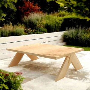 Puede incluir: Una mesa de centro de madera clara con una tapa rectangular y una base en forma de X única. La mesa está hecha de madera natural y tiene un estilo rústico y de granja.