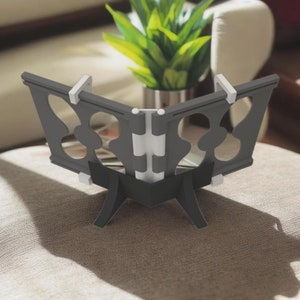 Puede incluir: Un soporte de Corán plegable negro con un diseño decorativo de corte y una bisagra blanca. El soporte tiene una base de tres patas.