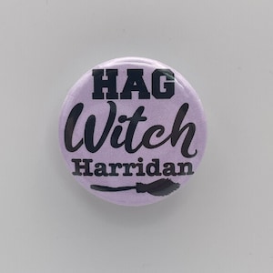 May include: A circular, pale purple button featuring the words "HAG Witch Harridan" in black. A black silhouette of a broom is at the bottom. This button is a novelty item, likely for a witch-themed product.