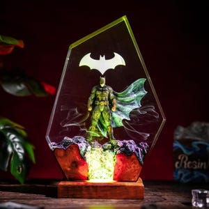 May include: A decorative resin display featuring a Batman figure encased in a clear, angular, crystal-like structure. The base is made of wood, with a glowing light illuminating the rocky terrain beneath the figure and a white bat symbol above.