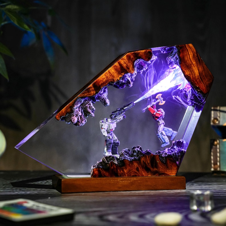 May include: A unique resin lamp featuring a battle scene with two action figures. The lamp has a wooden base and frame with a clear resin center. The figures are illuminated with purple and white light, creating a dramatic effect.