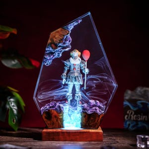 Könnte beinhalten: Eine dekorative Kunstharzskulptur mit einer Clownfigur, die einen roten Ballon hält, eingebettet in einen klaren, kantigen Kunstharzblock mit wirbelnden weißen Raucheffekten, auf einem rechteckigen Holzsockel mit blauer LED-Beleuchtung.