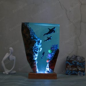 May include: A resin lamp with a blue and green ocean scene. The lamp features two orcas swimming in the water, a coral reef, and a jellyfish. The lamp is lit from the bottom, creating a glowing effect.