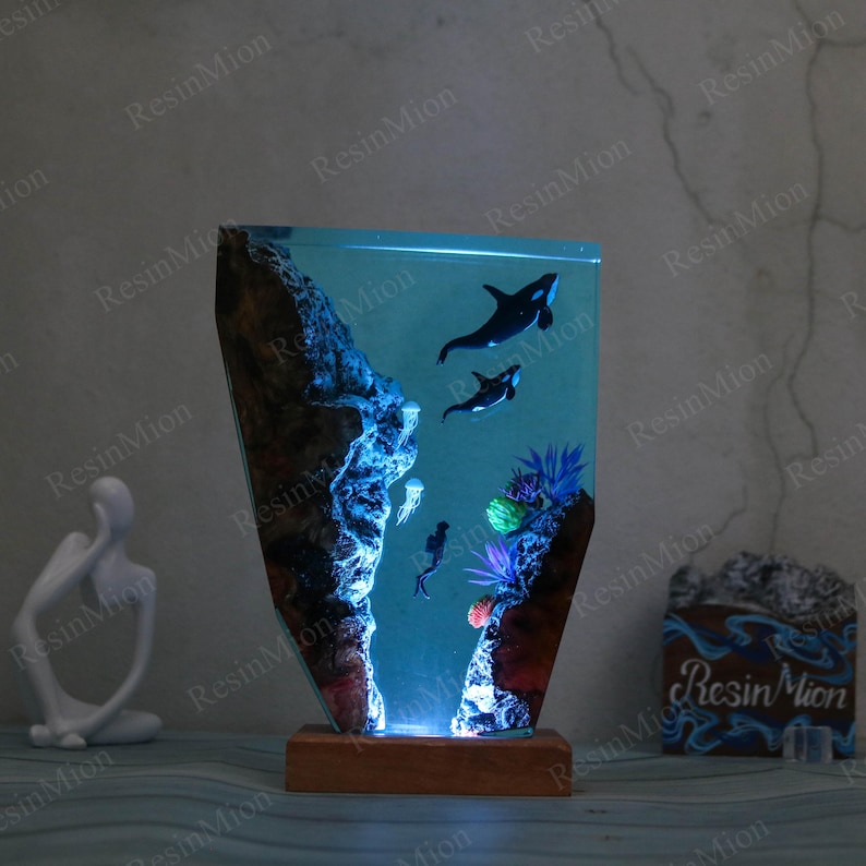 May include: A resin lamp with a blue and green ocean scene. The lamp features two orcas swimming in the water, a coral reef, and a jellyfish. The lamp is lit from the bottom, creating a glowing effect.