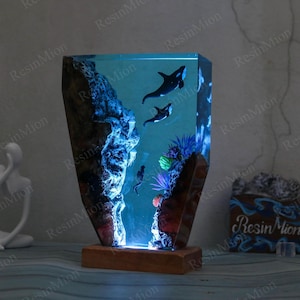 May include: A resin lamp with a blue and black ocean scene. The lamp features two orcas swimming in the water, coral, and a rock formation. The lamp is lit from the bottom, casting a blue glow.