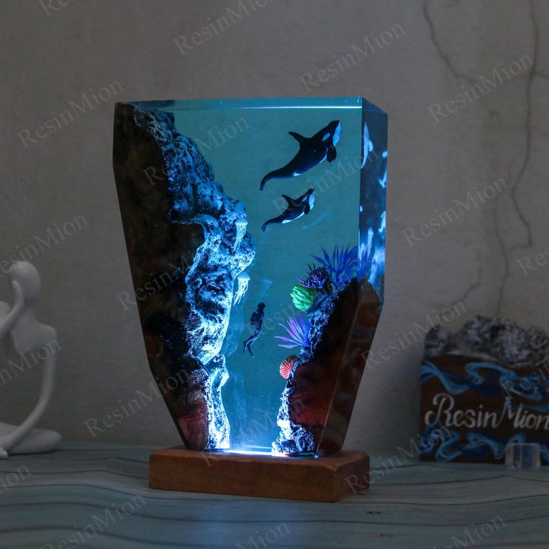May include: A resin lamp with a blue and black ocean scene. The lamp features two orcas swimming in the water, coral, and a rock formation. The lamp is lit from the bottom, casting a blue glow.