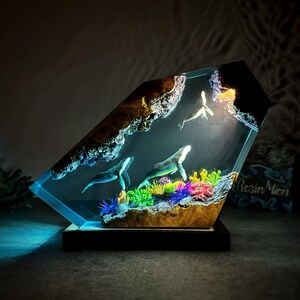 May include: A resin sculpture of three whales swimming in a blue and green ocean scene. The sculpture is lit from within, creating a glowing effect. The whales are made of clear resin and have a realistic appearance. The ocean scene is made of resin and includes coral, seaweed, and other marine life.