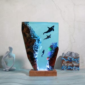 May include: A blue resin sculpture depicting an underwater scene with a diver, orcas, and coral. The sculpture is lit from below and sits on a wooden base. The base has the text "Resin Mion" written on it.