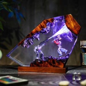 May include: A unique resin lamp featuring a battle scene with two action figures. The lamp has a wooden base and frame with a clear resin center. The figures are illuminated with a blue light, creating a dramatic effect. The lamp is a decorative item.