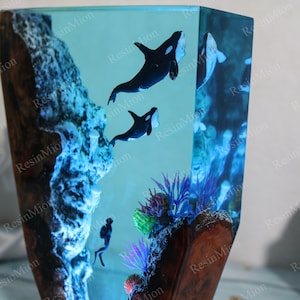 May include: A clear resin sculpture of an underwater scene with three black and white orcas swimming through a blue and green coral reef. The sculpture is set in a wooden base.