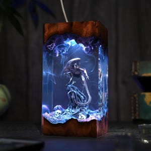 May include: A rectangular lamp featuring a dark wood frame and a clear resin center. Inside the resin, a silver-toned xenomorph figure stands atop a textured base with small skulls, illuminated by a blue light that creates a smoky effect.