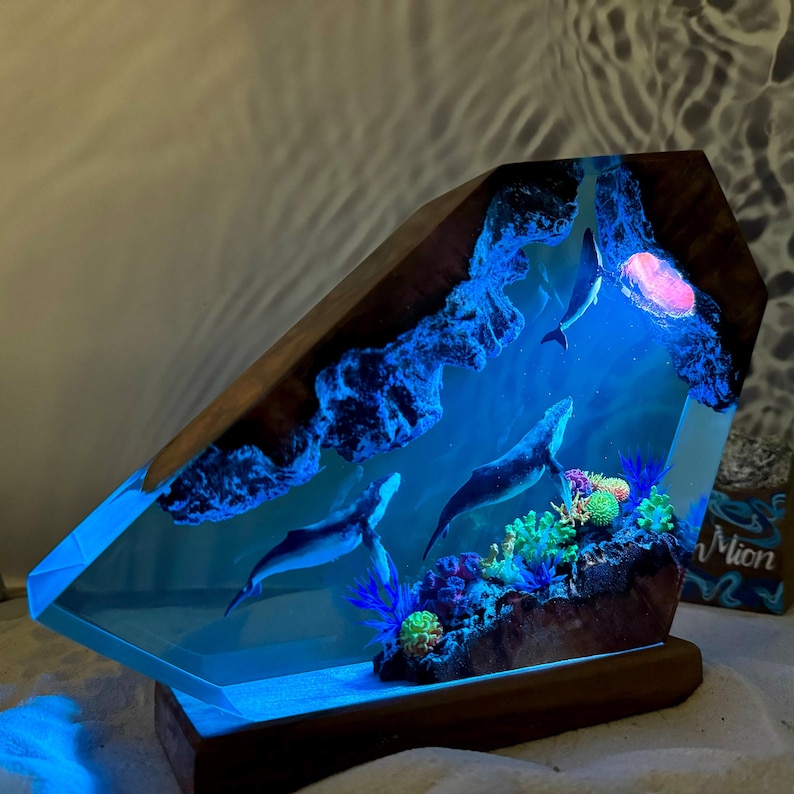 May include: A resin sculpture of a whale family swimming in a blue ocean with coral reefs. The sculpture is lit from within, creating a glowing effect. The sculpture is mounted on a wooden base.
