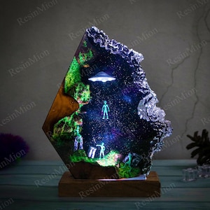 May include: A decorative resin art piece depicting an alien landscape with a UFO and figures, set against a starry backdrop. The artwork is presented in a geometric form, mounted on a wooden base, and illuminated with internal lighting.