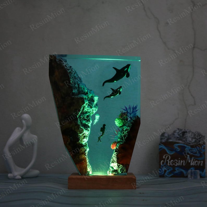 May include: A resin lamp with a green base and a clear resin section depicting an underwater scene with two orcas, coral, and jellyfish. The lamp is lit from the bottom, casting a warm glow.