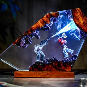 May include: A unique lamp featuring a battle scene with two robot figures encased in clear resin. The lamp has a wooden base and a geometric shape, with a blue light emanating from the center. The resin is bordered by a dark, textured material.