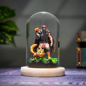 May include: A figurine of a muscular figure with a mask, leather vest, and cape, holding a flaming object, stands with a cat figurine under a glass dome. The figure wears heart-patterned underwear. The base is wood.