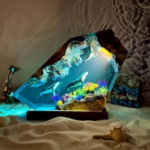 May include: A resin sculpture of a whale and coral reef, encased in a clear resin block. The sculpture is illuminated from within, creating a glowing effect. The base of the sculpture is made of wood.