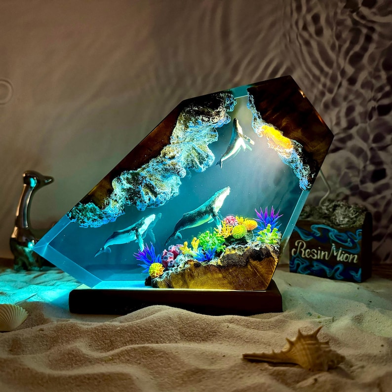 May include: A resin sculpture of a whale and coral reef, encased in a clear resin block. The sculpture is illuminated from within, creating a glowing effect. The base of the sculpture is made of wood.