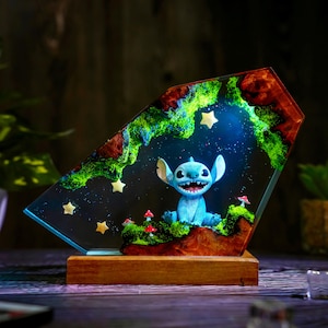 May include: A blue resin night light featuring a cartoon character with large ears and a wide grin, set against a starry night sky. The light is framed by a colorful landscape with green moss, red mushrooms, and yellow stars, mounted on a wooden base.