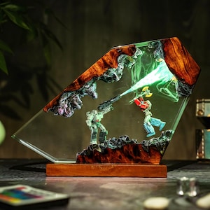 May include: A unique, illuminated resin art piece featuring a battle scene with two action figures. The artwork is encased in a clear, angled resin block with a wooden base and accents. The figures are in the middle of a battle, with a green light emanating from the top.