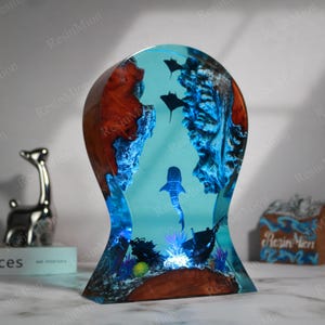 May include: A resin sculpture of a whale shark swimming in a blue ocean with coral and other fish. The sculpture is made of wood and resin and has a light blue base.