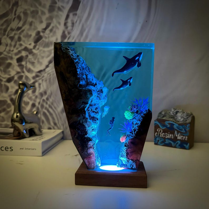 May include: A resin lamp with a blue and green ocean scene. The lamp features two orcas swimming in the water, coral, and a jellyfish. The lamp is lit from the bottom, creating a glowing effect.