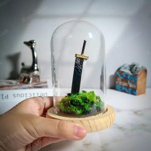 May include: A miniature black and gold sword replica, displayed under a glass dome. The sword rests on a bed of green moss and sits on a wooden base. The dome is held by a hand. Decorative items are in the background.