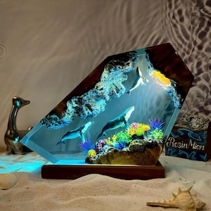 May include: A resin sculpture of three whales swimming in a blue ocean with coral reefs and a wooden base. The sculpture is lit from below, creating a glowing effect.