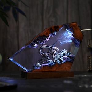 May include: A unique, illuminated resin sculpture featuring an alien creature within a wooden frame. The sculpture has a dark color palette with blue and purple lighting effects. The base is a dark brown color.