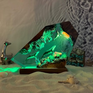 May include: A resin sculpture of three whales swimming in a green ocean scene. The sculpture is lit from within, creating a glowing effect. The base is made of wood and has the text "Resin Moon" on it.