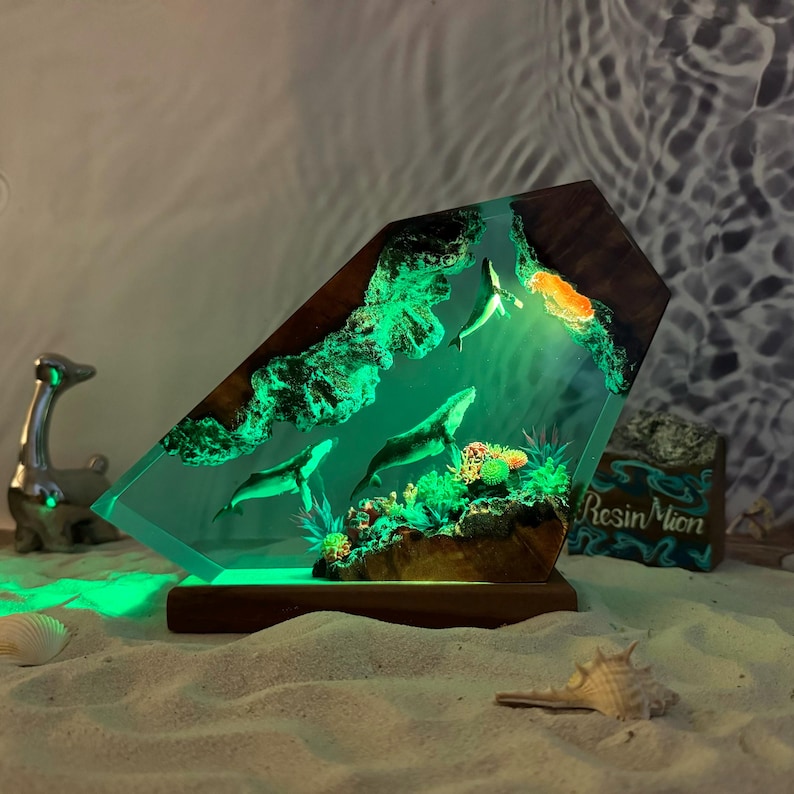 May include: A resin sculpture of three whales swimming in a green ocean scene. The sculpture is lit from within, creating a glowing effect. The base is made of wood and has the text "Resin Moon" on it.