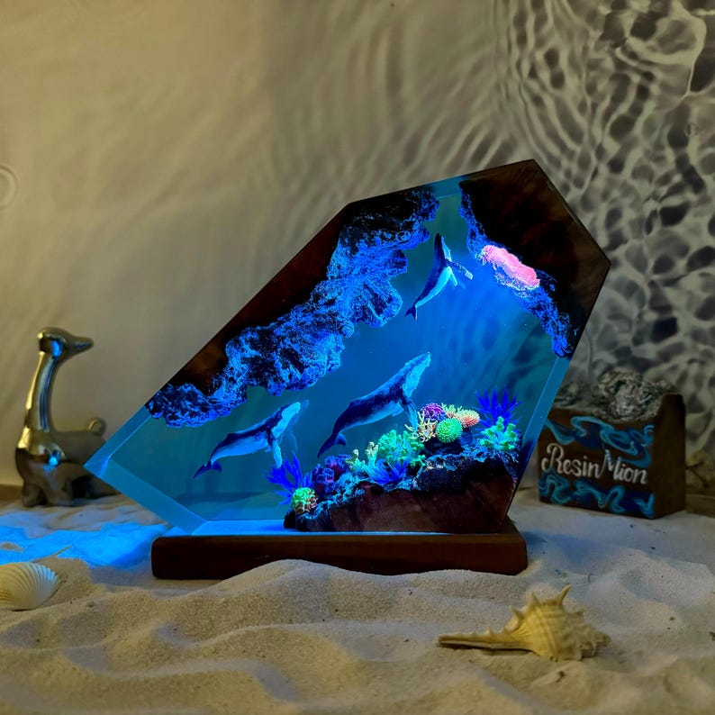 May include: A resin sculpture of three whales swimming in a blue resin ocean with coral and other sea life. The sculpture is lit from within, creating a glowing effect. The sculpture is mounted on a wooden base.