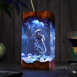 May include: A rectangular wooden lamp with an Alien figure encased in blue resin. The lamp features a detailed sculpture of the Alien, surrounded by blue, smoky effects. The wooden frame has a natural, dark brown grain.