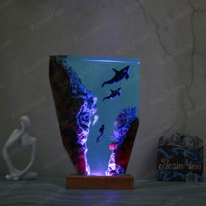 May include: A resin lamp with a blue and purple glow, featuring a scene of two orcas swimming in a coral reef. The lamp is on a wooden base and has a clear resin body.