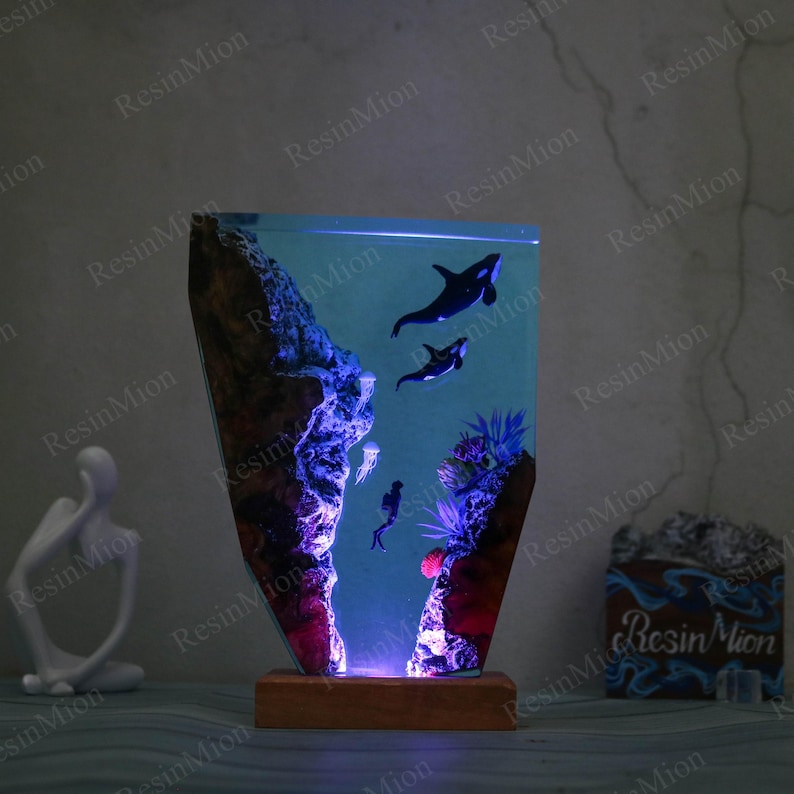 May include: A resin lamp with a blue and purple glow, featuring a scene of two orcas swimming in a coral reef. The lamp is on a wooden base and has a clear resin body.
