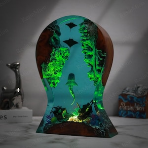 May include: A resin sculpture depicting a whale shark and manta rays swimming in a blue and green ocean scene. The sculpture is illuminated from below, creating a glowing effect. The base of the sculpture is made of wood.