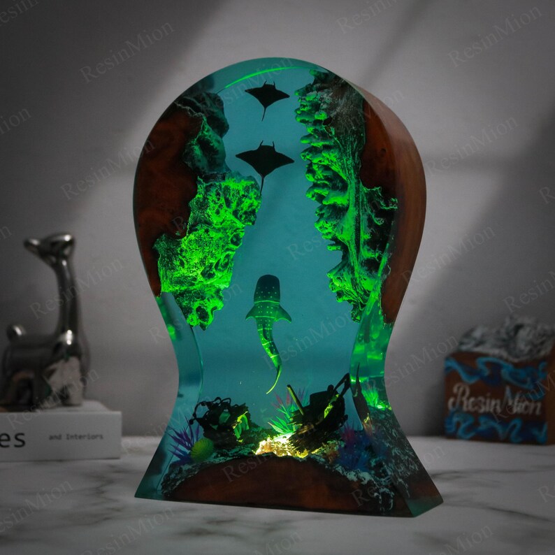 May include: A resin sculpture depicting a whale shark and manta rays swimming in a blue and green ocean scene. The sculpture is illuminated from below, creating a glowing effect. The base of the sculpture is made of wood.