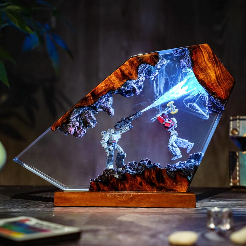 May include: A unique resin art piece featuring two robot figures in a battle scene. The artwork is encased in clear resin with a wooden frame and base. The figures are gray and red, with a blue light effect.