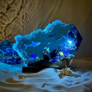 Könnte beinhalten: Eine blaue Harzskulptur, die eine Unterwasserszene mit einem Hai, einer Schildkröte und Korallen darstellt. Die Skulptur wird von hinten beleuchtet, wodurch ein leuchtender Effekt entsteht. Die Skulptur steht auf einem Holzsockel und befindet sich auf einer sandigen Oberfläche.