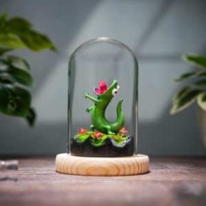 Può includere: Una stravagante statuina di lucertola verde con la lingua rosa, esposta sotto una campana di vetro. La lucertola è su una piccola base muschiosa con accenti di funghi rossi, posta su un supporto di legno. Un oggetto decorativo.