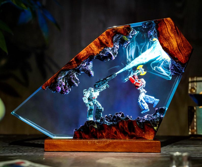 May include: A unique resin art piece featuring a battle scene between two robot figures. The artwork is encased in a clear, angled resin block with a wooden base. The figures are set against a blue and purple background with a beam of light.