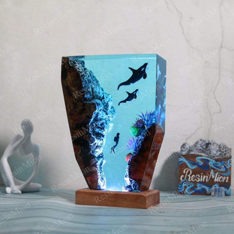 May include: A resin lamp with a blue and white ocean scene. The lamp features two orcas swimming through a coral reef. The base of the lamp is made of wood.
