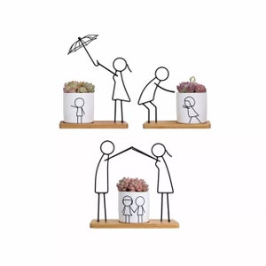May include: Three white ceramic planters with black stick figure designs. The planters are held by black metal stick figures. The figures are holding an umbrella, reaching for a plant, and holding hands over a plant. The planters are on wooden shelves.