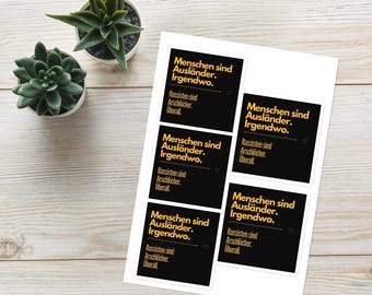 May include: A sheet of stickers with black rectangles and gold text. The text reads "People are foreigners somewhere. Racists are assholes everywhere." Two small potted succulents are in the background.