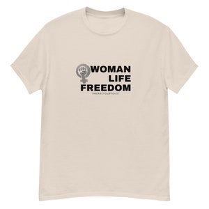 Könnte beinhalten: Ein beiges T-Shirt mit einem schwarzen grafischen Aufdruck, der "WOMAN LIFE FREEDOM" mit einem feministischen Symbol und einer Faust sagt. Der Text enthält auch den Hashtag #WEAREYOURVOICE.