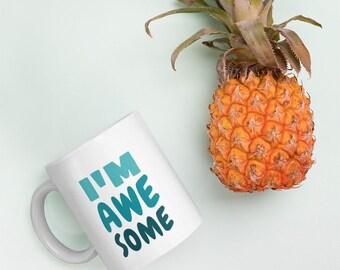 以下が含まれることがあります： 白地に青と緑のグラデーションで「I'M AWESOME」と書かれた白いセラミックマグカップ。マグカップは、薄い緑色の背景にパイナップルが置かれている。