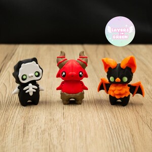 Op de afbeelding: Drie kleine, kleurrijke beeldjes: een zwarte Grim Reaper, een rode duivel en een zwarte vleermuis met oranje vleugels. De Grim Reaper heeft een wit schedelgezicht en een botontwerp. De duivel heeft hoorns. De vleermuis heeft groene ogen. Het Layers in Green-logo staat op de achtergrond.