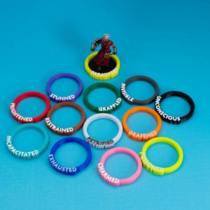 May include: A selection of brightly coloured, circular game tokens, each with white text denoting status effects such as "Stunned" and "Charmed." A miniature figure stands on a yellow token labelled "Paralyzed." The tokens are on a bright blue surface.